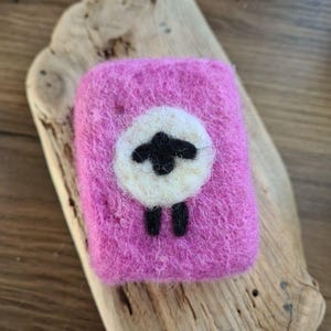 May include: A pink felted soap bar with a white sheep design. The sheep has black eyes and legs.