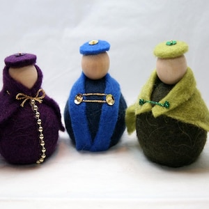 May include: Three handmade felt dolls with wooden heads. The dolls are dressed in purple, blue, and green felt outfits, each with a matching hat. The purple doll has a gold bow and beaded trim. The blue doll has a gold pin. The green doll has a green trim.