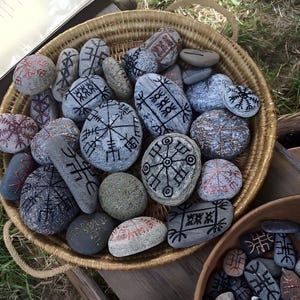 May include: A wicker basket overflowing with grey stones, each decorated with detailed black and red symbols. The stones are of different sizes and shapes, forming a unique collection. The basket sits on green grass.