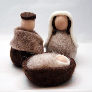 Puede incluir: Un belén artesanal con tres figuras de fieltro: José, María y el niño Jesús en un pesebre. Las figuras están hechas de lana marrón, beige y blanca. La escena se desarrolla sobre un fondo blanco liso.