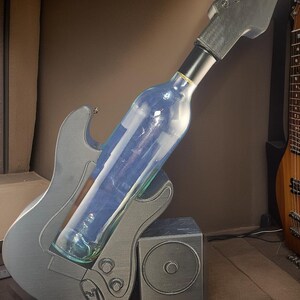 May include: A guitar-shaped wine bottle holder in silver with a black neck and headstock. A clear wine bottle is held in place. A small speaker-shaped stand is next to the guitar. The background is a neutral brown.