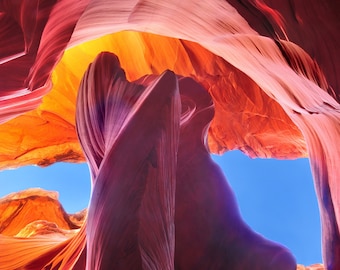 Könnte beinhalten: Ein Foto eines Sandstein-Canyons mit geschichteten Felsformationen in Rot-, Orange- und Lilatönen. Die Canyonwände sind geschwungen und verdreht und umrahmen einen strahlend blauen Himmel. Das natürliche Licht erzeugt einen dramatischen Effekt.