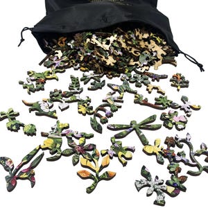 May include: Wooden jigsaw puzzle pieces with floral and leaf designs spill from a black drawstring bag. The pieces feature vibrant colors and intricate details, including flowers, leaves, and a dragonfly. The puzzle appears to be nature-themed.