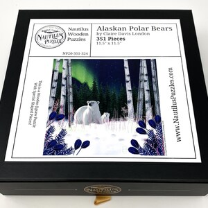 May include: A wooden jigsaw puzzle box featuring a snowy scene with two polar bears in a birch forest under a starry sky with a green aurora borealis. The box is black with a white label that reads "Alaskan Polar Bears by Claire Davis London 351 Pieces 11.5" x 11.5" NP20-351-324 This is a Wooden Jigsaw Puzzle with Special Shaped Pieces! www.NautilusPuzzles.com".
