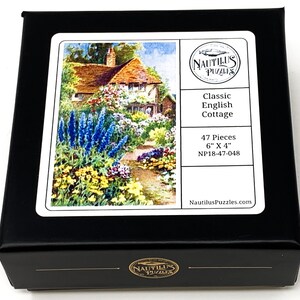 Puede incluir: Caja negra de rompecabezas con una imagen de una casa de campo inglesa cl&aacute;sica rodeada de flores. La caja est&aacute; etiquetada como "Classic English Cottage", con 47 piezas y mide 15 cm x 10 cm. La marca "Nautilus Puzzles" tambi&eacute;n es visible.