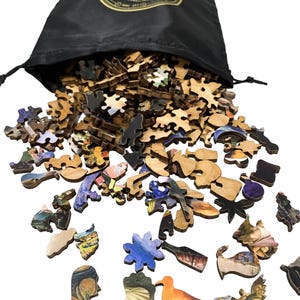 May include: A black drawstring bag with a gold logo spills out wooden jigsaw puzzle pieces. The pieces are various shapes and sizes, with colorful images of art and landscapes. The puzzle pieces are scattered on a white surface.