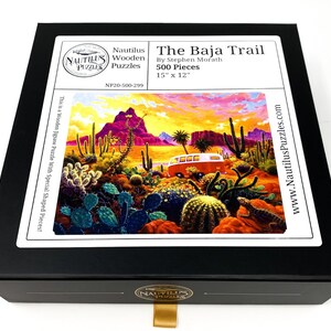 May include: A black box containing a jigsaw puzzle titled "The Baja Trail" by Stephen Morath. The puzzle features a desert landscape with a red van, cacti, and a lizard. The puzzle is 15" x 12" and has 500 pieces.