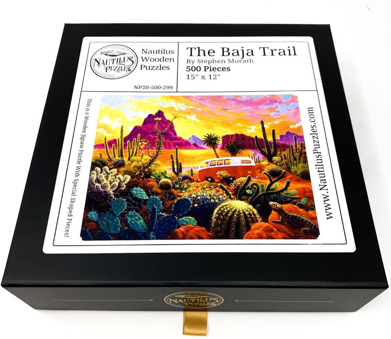 May include: A black box containing a jigsaw puzzle titled "The Baja Trail" by Stephen Morath. The puzzle features a desert landscape with a red van, cacti, and a lizard. The puzzle is 15" x 12" and has 500 pieces.