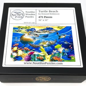 May include: A black box featuring a jigsaw puzzle titled "Turtle Beach" by Howard Robinson. The puzzle image depicts sea turtles, colorful fish, and coral in an underwater scene. The puzzle has 475 pieces and measures 16" x 11".