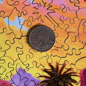 May include: A quarter coin rests on a colorful jigsaw puzzle. The puzzle features an abstract design with warm tones of yellow, orange, and purple, with black outlines. The puzzle's design includes shapes resembling palm trees and starbursts.