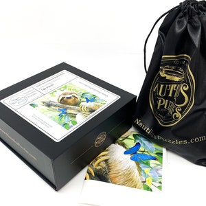 May include: A black box set of a wooden puzzle with the image of a sloth and butterflies. The box has a white label with the text "Sloth Among the Butterflies" and "143 Pieces". A black drawstring bag with gold text and logo is also in the image.