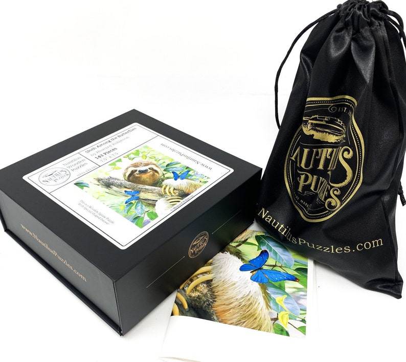 May include: A black box set of a wooden puzzle with the image of a sloth and butterflies. The box has a white label with the text "Sloth Among the Butterflies" and "143 Pieces". A black drawstring bag with gold text and logo is also in the image.
