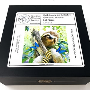 May include: A black box featuring a jigsaw puzzle with a sloth among butterflies. The puzzle, titled "Sloth Among the Butterflies" by Howard Robinson, contains 143 pieces and measures 7.5" x 7.5". The box also displays the Nautilus Puzzles logo.