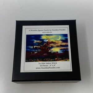 Może przedstawiać: Czarne pudełko z drewnianymi puzzlami przedstawiającymi wiedźmę lecącą na miotle nad miastem pod p&oacute;łksiężycem. Puzzle noszą tytuł "Ye Olde Salem Witch" i mają wymiary 15 cm x 10 cm.