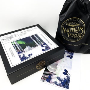 May include: A black box with a white label featuring a 351-piece jigsaw puzzle of two polar bears in a snowy forest with the aurora borealis in the background. The box is labeled "Nautilus Wooden Puzzles" and "Alaskan Polar Bears". The box is sitting on a white surface next to a black satin bag with the Nautilus Puzzles logo and a smaller image of the puzzle.