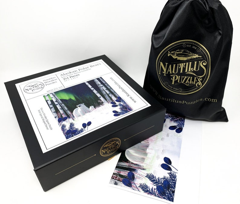 May include: A black box with a white label featuring a 351-piece jigsaw puzzle of two polar bears in a snowy forest with the aurora borealis in the background. The box is labeled "Nautilus Wooden Puzzles" and "Alaskan Polar Bears". The box is sitting on a white surface next to a black satin bag with the Nautilus Puzzles logo and a smaller image of the puzzle.