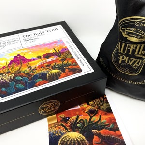 May include: A black box puzzle titled "The Baja Trail" by Nautilus Puzzles, featuring a desert landscape with cacti and a bus. The puzzle is 15 x 12 inches and includes 500 pieces. A black drawstring bag with the Nautilus Puzzles logo is also visible.