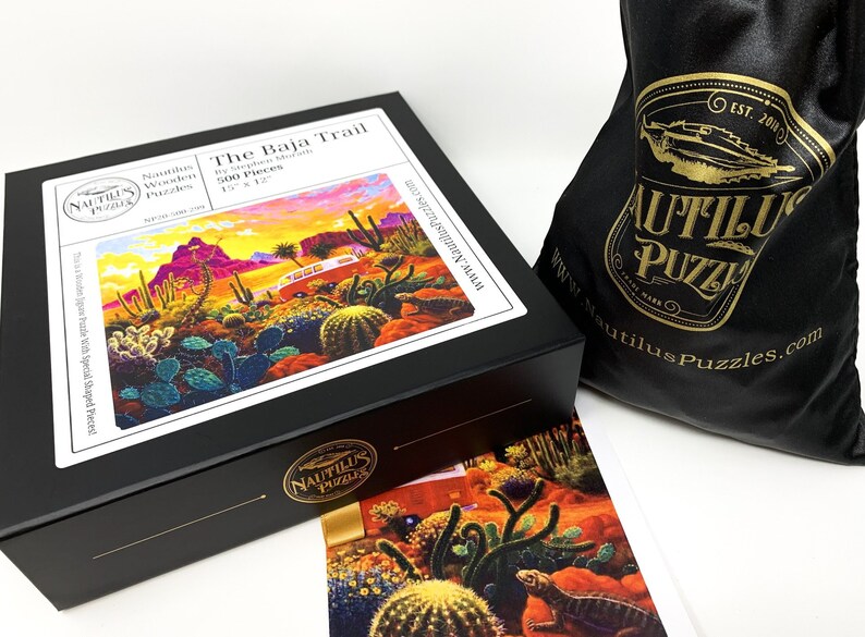 May include: A black box puzzle titled "The Baja Trail" by Nautilus Puzzles, featuring a desert landscape with cacti and a bus. The puzzle is 15 x 12 inches and includes 500 pieces. A black drawstring bag with the Nautilus Puzzles logo is also visible.