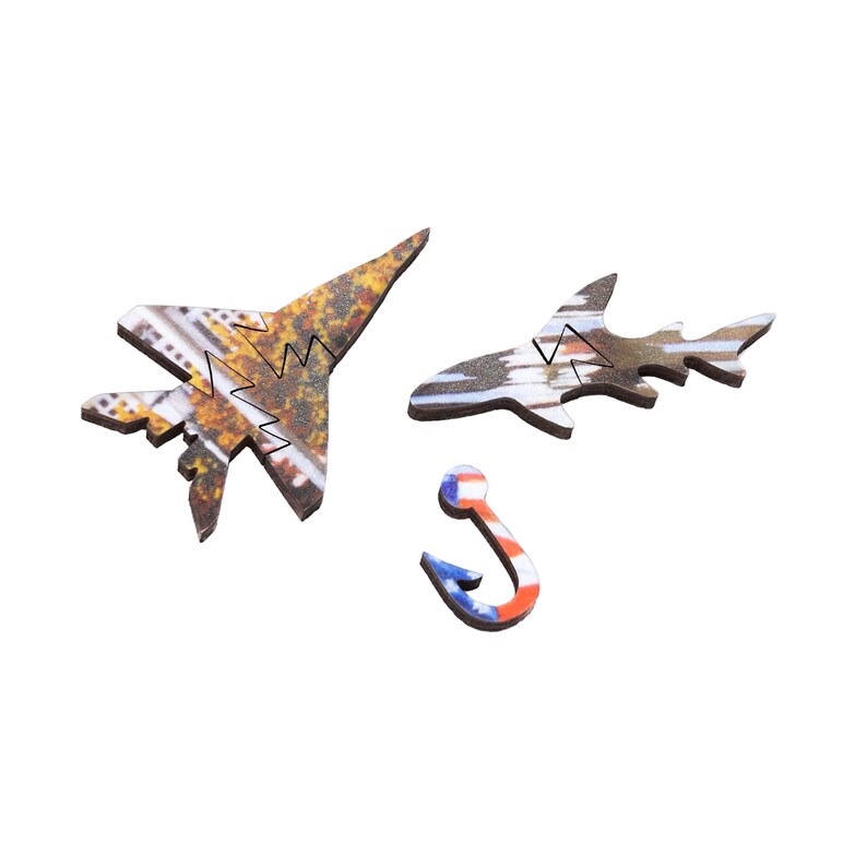 May include: Wooden puzzle pieces in the shapes of a jet, a shark, and a fishing hook. The jet and shark have a speckled, camouflage-like pattern. The hook is painted with red, white, and blue stripes and stars.