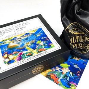 May include: A black box with a puzzle titled "Turtle Beach" by Howard Robinson. The puzzle is 16" x 11" and features an underwater scene with sea turtles and colorful fish. A black drawstring bag with a gold logo is next to the box.