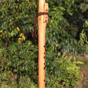 May include: A handcrafted wooden flute with a natural finish. The instrument features a series of finger holes and a decorative carved element near the mouthpiece. A brown cord wraps around the flute. The background is a blurred view of green foliage.