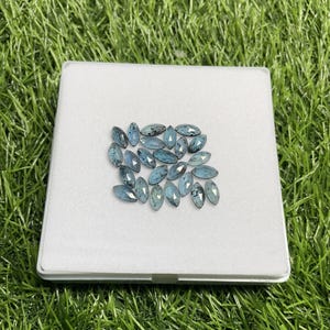 May include: A collection of marquise-cut gemstones in varying shades of blue and teal, displayed on a white surface. The gemstones are arranged in a cluster, showcasing their polished facets and internal patterns. The background is a vibrant green.