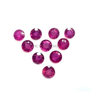 May include: A collection of ten round, faceted, deep pink-red gemstones arranged on a white background. The gemstones are evenly spaced and appear to be rubies. The text "THE BLUE GEMS" is visible in the image.