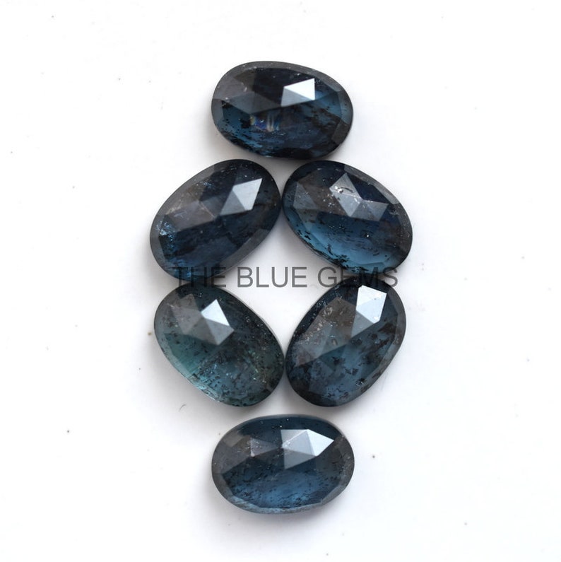 May include: Six dark blue faceted oval gemstones. The gemstones are arranged in a cluster on a white background.