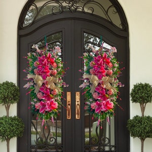 May include: Two floral door swags with vibrant pink flowers, green foliage, and gold ribbon bows adorn a black double door. The door has a decorative ironwork transom and brass hardware. Two topiary trees in bronze pots flank the door.