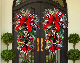 May include: Two Christmas door swags with a festive design. Each swag features a red bow, black and white checkered ribbon, and a nutcracker figurine. The swags are adorned with greenery and red berries, creating a holiday theme.