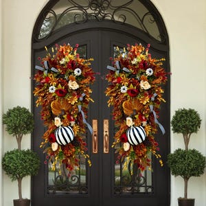 May include: Two fall-themed door swags with a mix of autumn leaves, flowers, and pumpkins. Each swag features a black and white striped pumpkin, red and cream-colored flowers, and decorative ribbons. The swags are displayed on a dark brown double door.