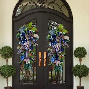 May include: Two matching door wreaths with patriotic themes. Each wreath features blue roses, red berries, and greenery, accented with ribbons in red, white, and blue. A banner with the word "WELCOME" is visible on each wreath.