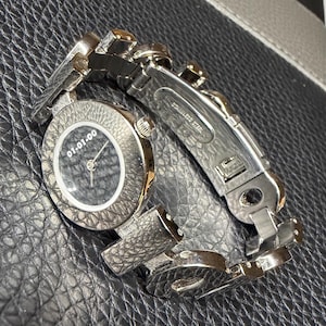 Puede incluir: Un reloj de pulsera plateado con una esfera circular gris oscuro texturizada que presenta el texto blanco 01-01-00. El reloj tiene un bisel texturizado a juego y una pulsera de eslabones de acero inoxidable pulido con un cierre marcado como stainless steel.