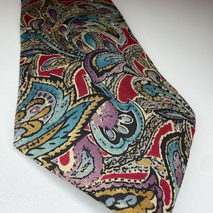 May include: A necktie featuring a complex paisley pattern in shades of red, teal, purple, gold, and black. The fabric has a visible ribbed texture throughout the design, which consists of swirling, organic shapes and intricate floral-inspired motifs.