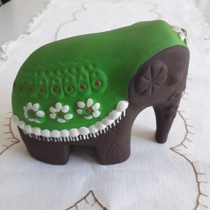 Puede incluir: Una figurita de elefante de cerámica marrón con un diseño pintado en verde y blanco. El elefante lleva un paño estampado en verde y blanco sobre su espalda.