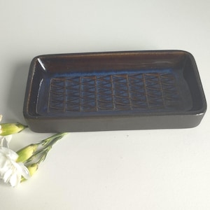 May include: A rectangular ceramic tray with a dark brown exterior and a glossy interior featuring a deep blue glaze. The base of the tray is decorated with a repeating embossed pattern of small triangles arranged in a grid formation.