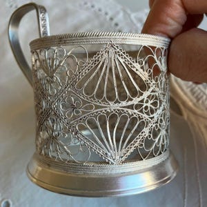 Könnte beinhalten: Ein silberfarbener Tee-Glashalter aus Metall mit einem komplexen Filigran-Design aus sich wiederholenden Rauten- und Fächermustern. Der zylindrische Rahmen verfügt über einen festen Boden und einen geschwungenen Griff mit feiner Drahtarbeit.