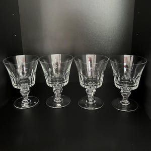 Op de afbeelding: Een set van vier heldere glazen bekers met een brede, uitlopende kom met verticale paneelsneden langs het onderste gedeelte, ondersteund door een gefacetteerde, meerzijdige steel en een ronde voet. Het glaswerk heeft een transparante afwerking.