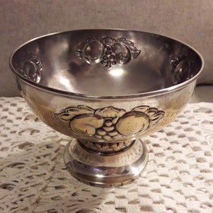 May include: A silver bowl with a pedestal base. The bowl has an embossed design of grapes and leaves around the rim and base.