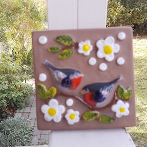 Puede incluir: Un azulejo de cerámica marrón con un diseño de mosaico que presenta dos pájaros con pechos rojos, flores blancas y hojas verdes. El azulejo tiene una superficie texturizada y un fondo blanco.