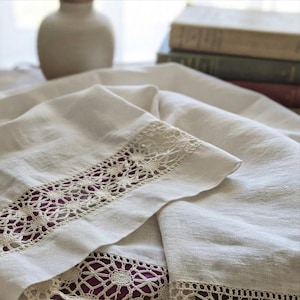 Può includere: Un tessuto bianco caratterizzato da un bordo in pizzo all'uncinetto color crema con un motivo circolare ripetuto. Il tessuto presenta una trama visibile ed è piegato per mostrare il bordo in pizzo dettagliato su una base di colore viola scuro.