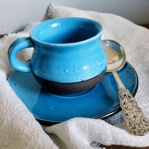 Puede incluir: Un juego de taza y plato de cerámica azul brillante con una base de color marrón oscuro. La taza presenta un asa redondeada y un acabado liso y brillante. Junto a la taza descansa una cuchara de metal plateado con un mango ornamentado.