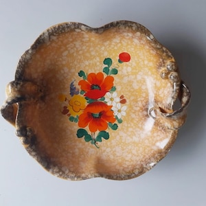 Op de afbeelding: Een vintage keramische serveerschaal met een gespikkelde gele en bruine buitenkant. De schaal heeft een bloemenontwerp met rode, oranje, gele en witte bloemen. Het heeft twee handvatten en een geschulpte rand.