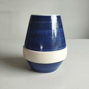 Könnte beinhalten: Eine Keramikvase mit konischer Silhouette und glänzender Oberfläche. Die oberen und unteren Bereiche sind in Dunkelblau mit horizontalen Pinselstrich-Texturen gehalten, getrennt durch ein breites, glattes, mattes cremeweißes Band in der Mitte.