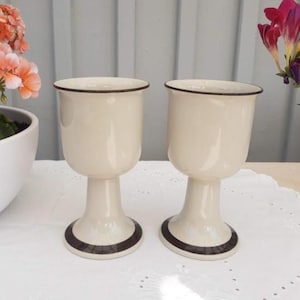 May include: Two off-white ceramic goblets with a smooth, glossy finish. Each goblet features a dark brown rim at the top and a matching dark brown circular band around the base. The stems are thick and cylindrical, supporting a rounded bowl shape.