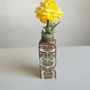 Peut inclure: Un vase carré en céramique avec un vernis à motifs marron, blanc et bleu sarcelle présentant des motifs géométriques abstraits. Le vase a un col étroit et une finition texturée, contenant une fleur artificielle jaune vif avec des feuilles vertes.