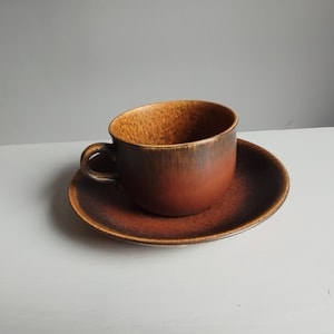 Puede incluir: Un juego de taza de té y platillo de cerámica con un esmalte de color marrón óxido y naranja quemado con una textura moteada y salpicada. La taza tiene un cuerpo redondeado y un asa curva, mientras que el platillo tiene un borde ancho.
