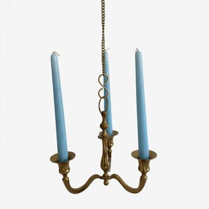 Peut inclure: Un candélabre suspendu en laiton doté de trois bras, chacun supportant une longue bougie effilée bleu clair. Le luminaire est suspendu par une fine chaîne en laiton reliée à un montant central vertical, formant un design équilibré à trois lumières.