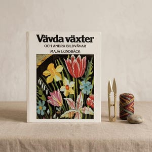 Könnte beinhalten: Ein weißes Hardcover-Buch mit dem Titel Vävda växter och andra bildvävar von Maja Lundbäck, das auf dem Einband eine florale Wandteppich-Illustration zeigt. Neben dem Buch liegen eine goldfarbene Metall-Fadenschere, eine Spule mit mehrfarbigem Garn und ein kleiner grauer Stein.