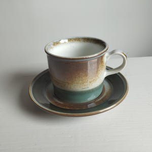 May include: A ceramic cup and saucer set featuring a gradient glaze. The cup has a white interior, a brown upper band, and a teal lower band. The saucer is dark gray with a brown rim, matching the earthy tones of the cup's exterior design.